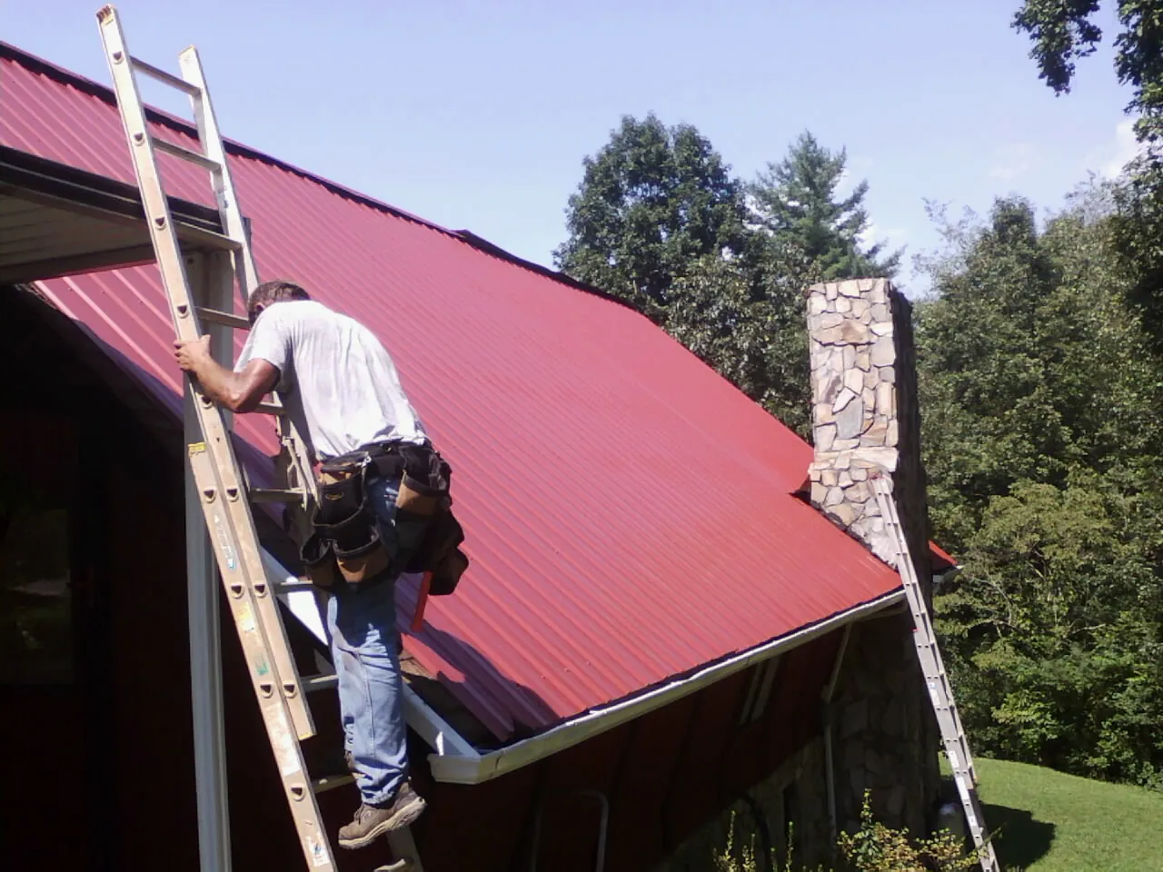 Roofer on a ladder beside a red metal roof and stone chimney — Soffit & Fascia Repair by Roofing Brothers in North Troy